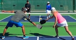 Four adults playing pickleball on a court. Two players are at the net, with one man in a gray shirt and blue shorts reaching for a ball with a paddle, while a woman in a pink tank top prepares to hit the ball.