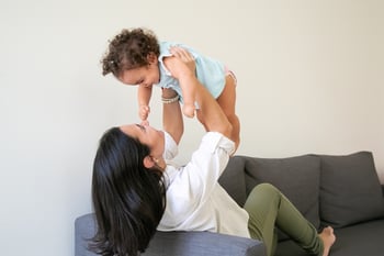 happy-mother-holding-baby-girl-arms-lifting-child-while-sitting-couch-home-parenthood-childhood-concept
